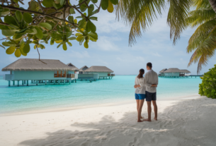 Beautiful Places to Travel: A couple arm in arm on the beach looking over the beautiful aqua water to the beautiful overwater bungalows on a beach in the Maldives