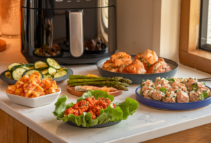 A selection of low carb snacks and lunches that can be made in an air fryer. Show the air fryer and dishes of Buffalo Chicken Lettuce Wraps, Air Fryer Zucchini Chips, Garlic Parmesan Asparagus, Air Fryer Herbed Chicken Thighs and Tuna Salad Bites on a cream colored marble bench in an inviting kitchen atmosphere.