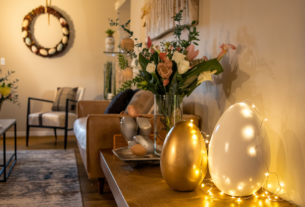 A warm modern living room elegantly decorated for Easter with fairy lights, gold and white egg sculpture, floral arrangements and pine cone and egg wreath.